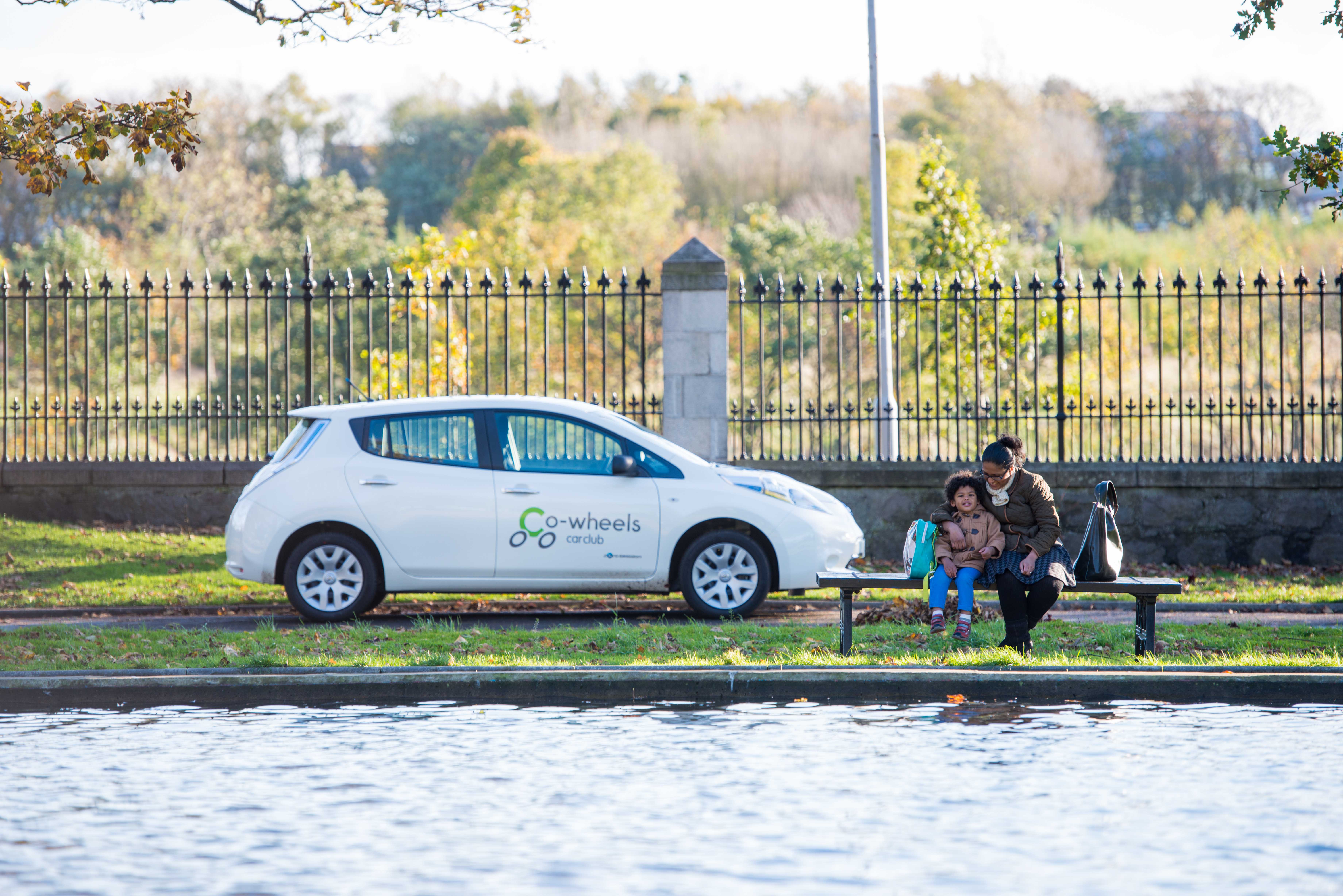 A photo of a mother and child sitting on a bench by a pond, with a Co-Wheels shared car in the background.