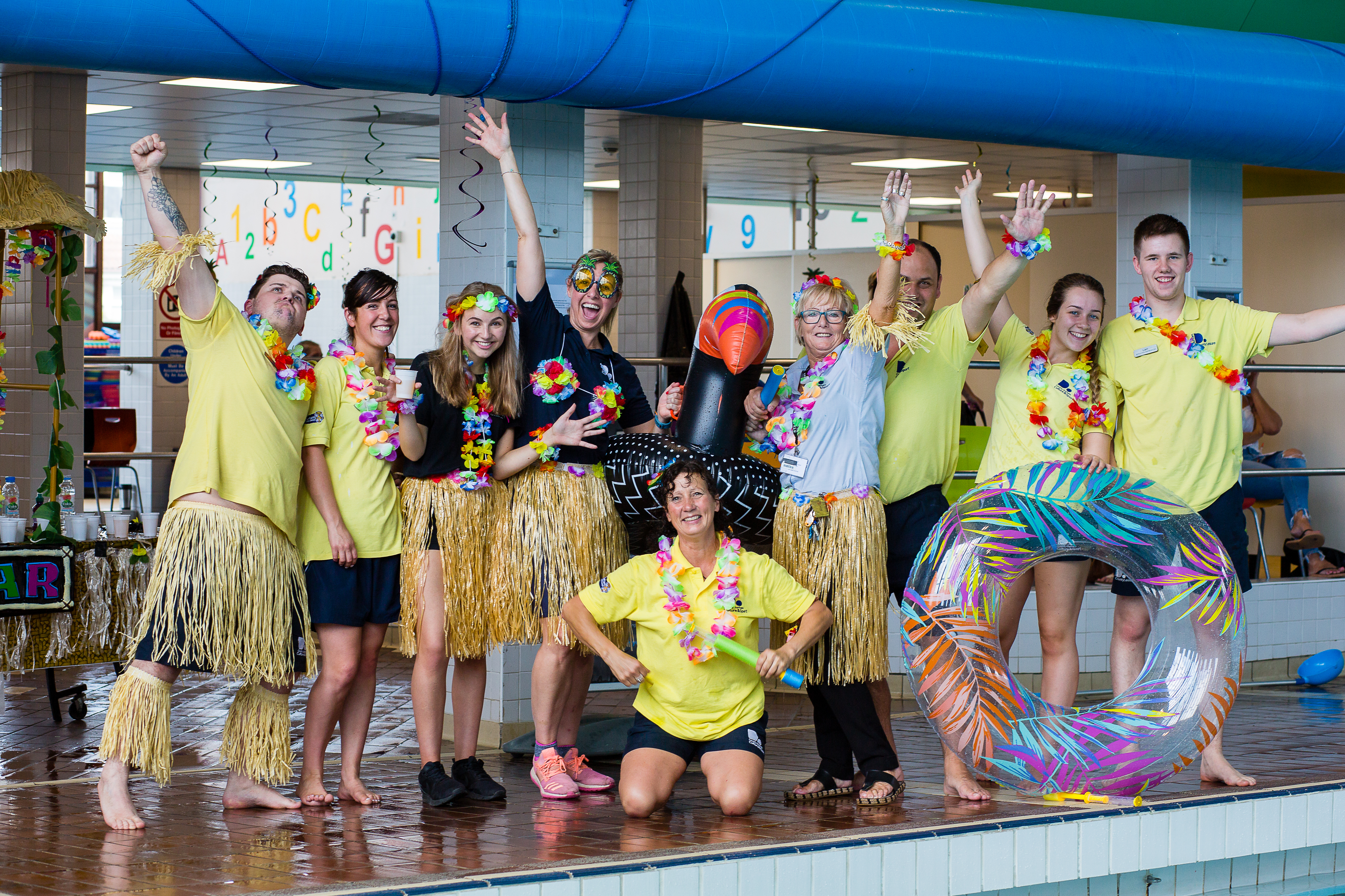 A group of leisure centre staff, dressed in grass skirts and flower garlands, stand and kneel at the side of a pool ready for the start of a themed pool party.
