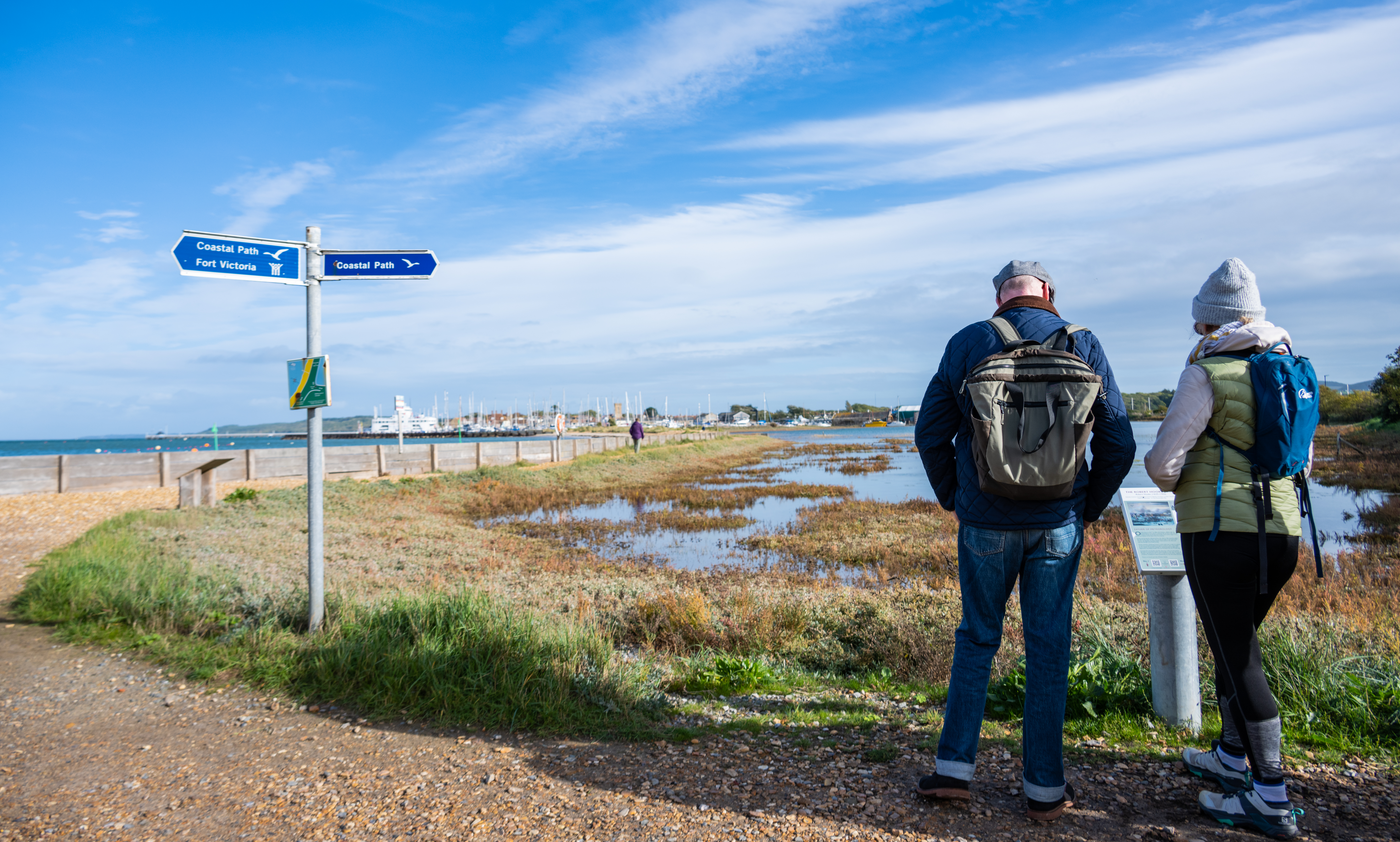 Photograph of a middle aged white man and woman looking at an interpretation board near a signpost on the Isle of Wight coastal path.
